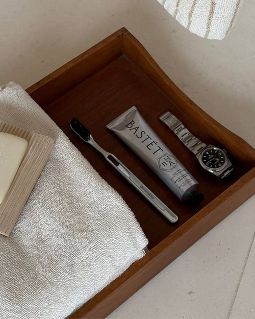 Bastét aluminium toothpaste tube in a wooden tray with a toothbrush, a watch and a folded towel